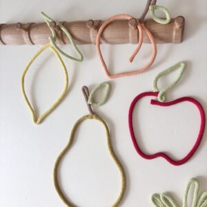 Set of six handmade fruit wall decorations including lemon, pear, peach, apple, pineapple and cherries displayed on a wooden peg rail in a colourful kids room.