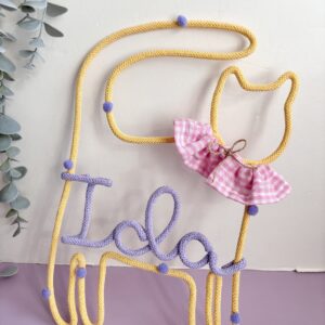 Personalised butter-yellow cat wall decoration with lilac name, lilac pom-poms and a checkered collar for a child's bedroom.